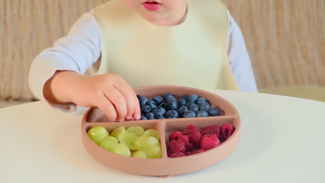 The child happily eats berries and fruits while sitting at the table in their beige home. Kid boy aged two years (two-year-old)