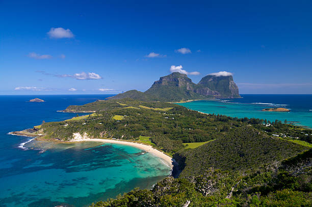 View south over the stunningly beautiful Lord Howe Island from Malabar lookout. Lagoons on both sides of the island are visible, as well as the peaks of Mts Lidgbird and Gower at the far end of the island. The sky and ocean are brilliant blue.