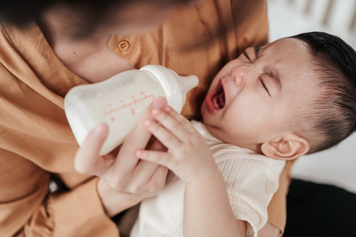 Adorable little Asian baby uncomfortable crying during drinking from baby bottle. Mother holding adorable her son with milk feeding from milk bottle. Concept babies healthcare.