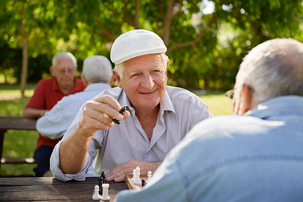 Senior men playing chess in a park with blurred men in back Active retired people, old friends and free time, two seniors having fun and playing chess game at park. Waist up senior chess stock pictures, royalty-free photos & images