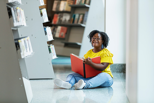 A multiracial teenage girl, 15 years old, sitting on the floor in a library, reading a book. She has down syndrome. She is looking at the camera, laughing.