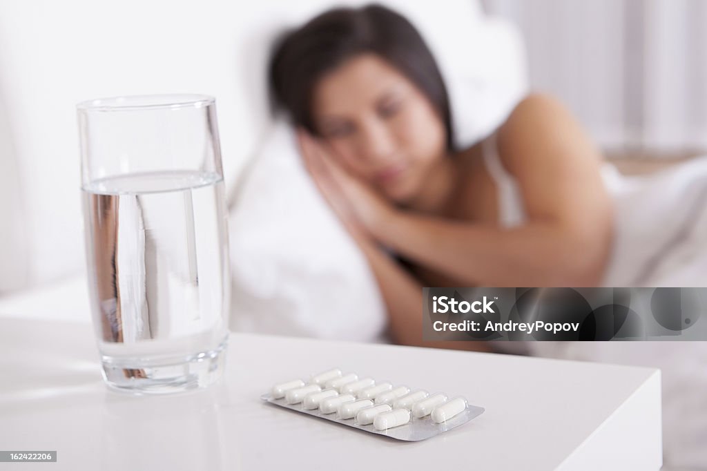 Sick woman resting in bed Sick woman resting in bed with focus to a glass of water and a blister pack of tablets and medication lying on the table alongside her bed Adult Stock Photo