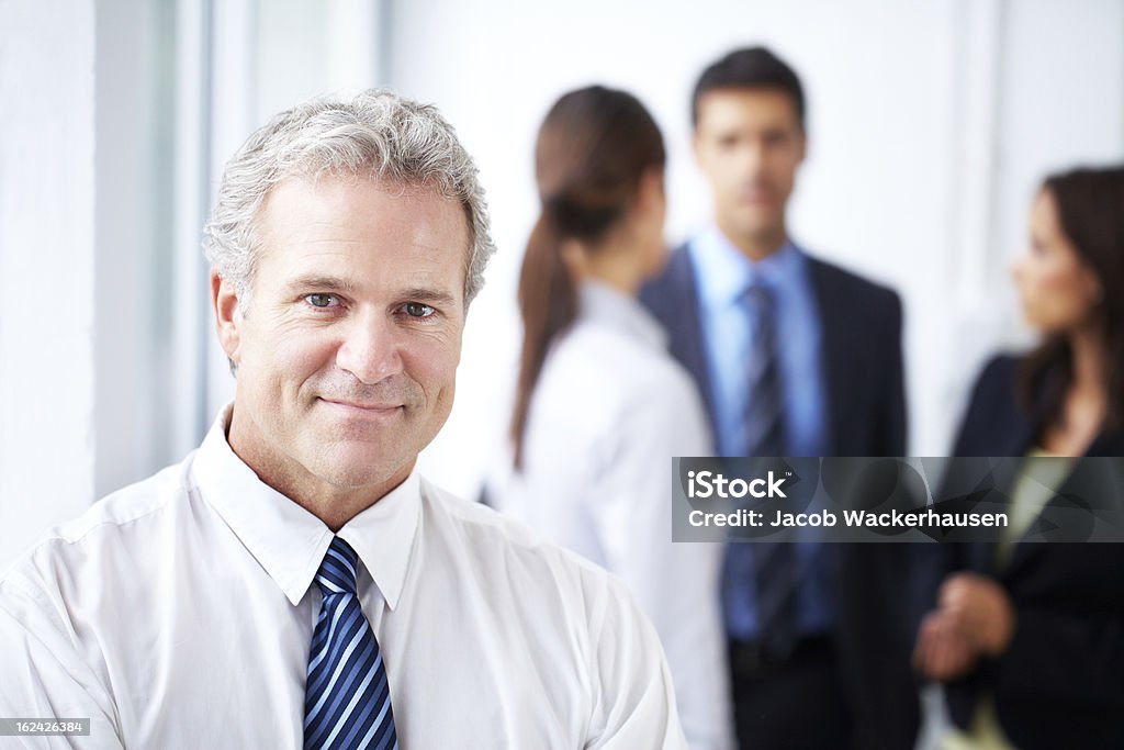 Creating a pleasant work environment is paramount Happy mature businessman smiling at the camera while his colleagues are standing in the background Adult Stock Photo