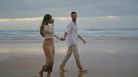 Young spouses relaxing beach at evening. Happy lovers holding hands walking together at sea shore. Loving woman man having romantic honeymoon date at dusky ocean side. Love relationship concept