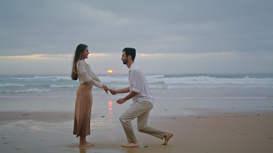 Amorous lovers engaging at sunset seashore. Handsome man making surprise marriage proposal to beloved woman at evening beach. Rejoicing girl saying yes. Emotional moment. Relationship goals concept