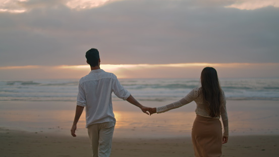 Young spouses relaxing sunbeams beach at cloudy evening. Happy lovers holding hands walking together at sea coast back view. Loving woman man romantic honeymoon date at ocean side. Love relationship