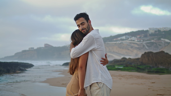 Enamoured man carrying wife at marine landscape closeup. Beautiful couple enjoying sea beach. Happy sweethearts hugging together at cloudy day. Romantic lovers embracing at ocean side. Love concept