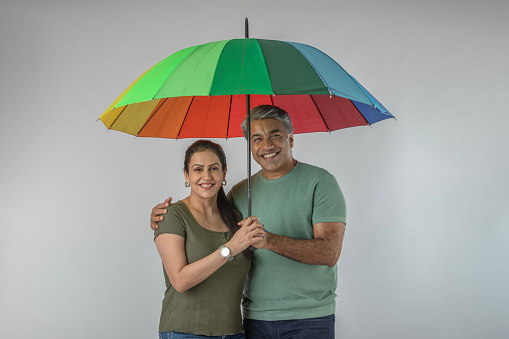 Portrait of smiling Indian husband and wife holding multicolored umbrella while standing against white background