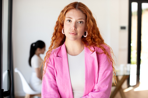Portrait of beautiful redhead business woman or office worker. A young and dynamic professional stands in a white-walled room. She exudes seriousness, wearing a pink blazer. In the background her colleagues are working at the table.