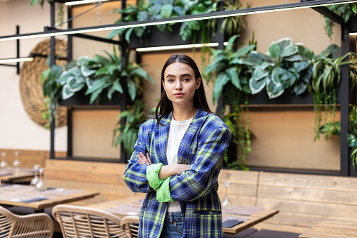 Portrait of beautiful young business woman, restaurant owner or manager. A young and promising professional or trainee stands in a room with green plants in the background. She exudes a serious demeanor, dressed in a checked blazer, hands folded on her chest, in a business posture.