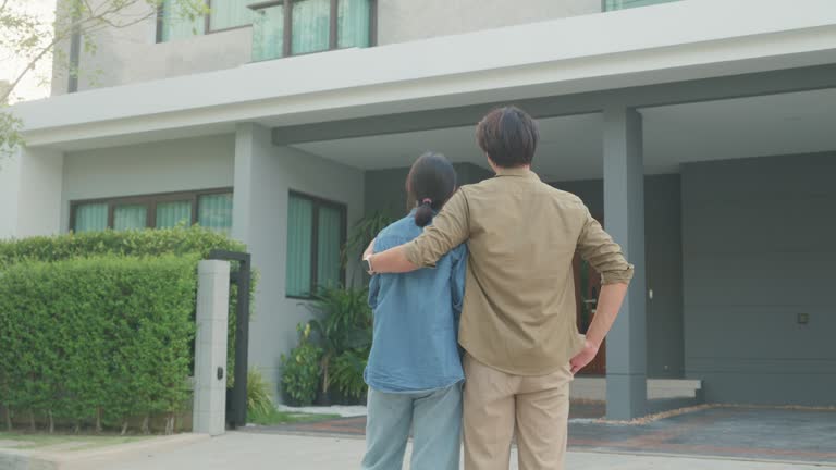 Young Asian couple standing and hugging together looking happy in front of their new house. Buying a new house.