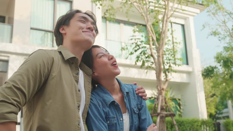 Young Asian couple standing and hugging together looking happy in front of their new house. Buying a new house.