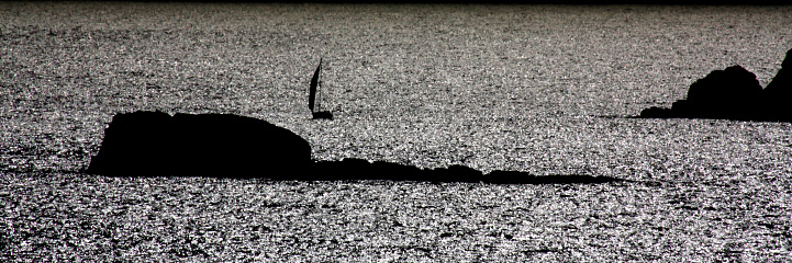 Sailboat silhouette and small islands back lit, waves and coastline at dusk. Rías Baixas, Galicia, Spain.