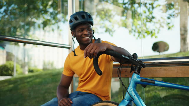 Positive African American man sits at bus stop with bicycle
