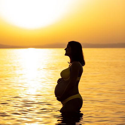 Photo of a eight month pregnant woman with hands on her abdomen at the seaside in sunrise