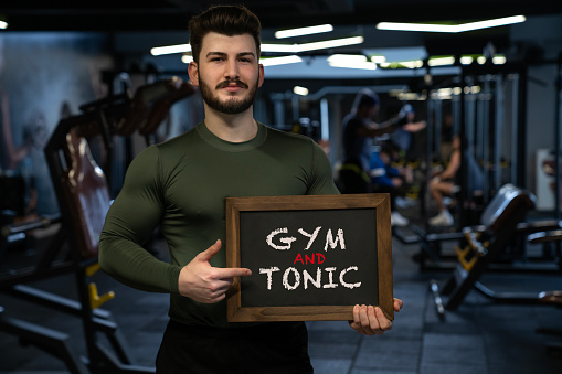 A profile view of a powerful young adult man at the fitness center. Gym and Tonic. Handsome young sporty sexy man showing a blackboard.