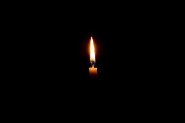 istock Single burning candle flame or light glowing on a small white candle on black or dark background on table in church for Christmas, funeral or memorial service with copy space. 1625006546