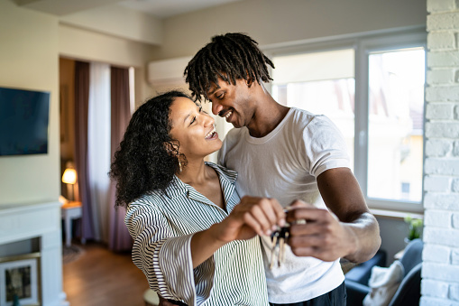 A young couple are happily holding the keys to their first home together as they share an embrace, smiling happily at each other standing in the furnished modern living room