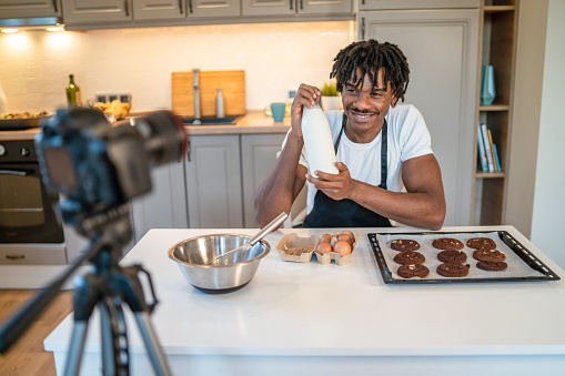 Using a digital camera to record a e-learning video a man is holding a bottle of milk up while leaning on the kitchen counter, covered in cookies and eggs and a mixing bowl