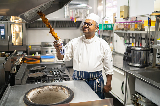 In a large industrial restaurant kitchen, an Indian chef is looking at a chicken kebab he has taken from the tandoori oven in front of him, behind the rest of the kitchen can be seen
