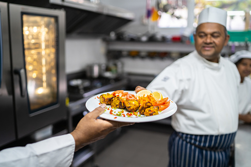 A smiling chef is passing a plate of garnished spicy Indian food to another hand in a large restaurant kitchen