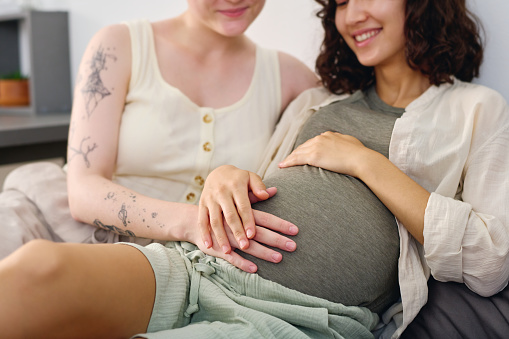 Focus on hands of two girlfriends touching pregnant belly of one of them while relaxing on bed in front of camera and enjoying being together
