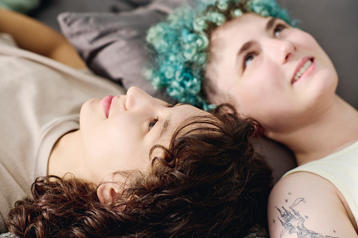 Focus on face of young serene brunette woman lying on bed in front of camera against her smiling girlfriend with short blue curly hair