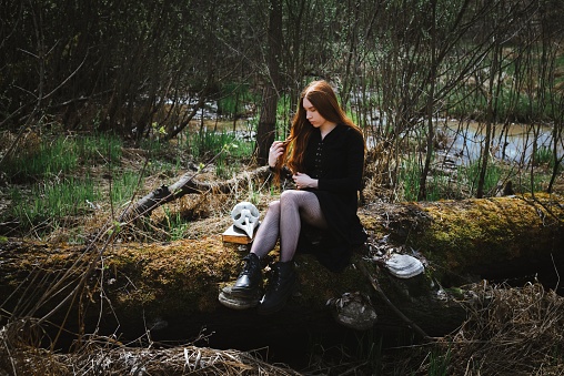 Woman sitting in a black robe with a skull and book in the dark forest swamp. Mystical portrait of a witch. The concept of witchcraft and occultism.