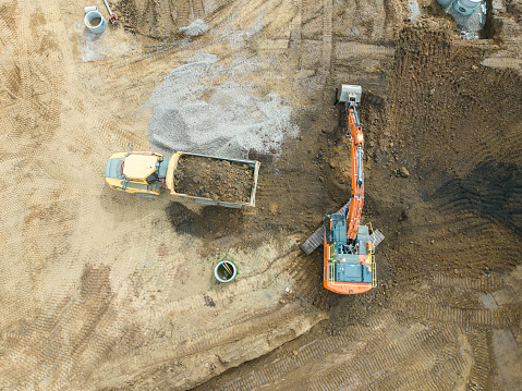Aerial drone view of a housing development construction site in Scotland.  A bulldozer is at work, levelling the ground, with soil being taken away in a dumper truck, so the site will be ready for the building of new houses to start. The heavy machinery is seen directly below.