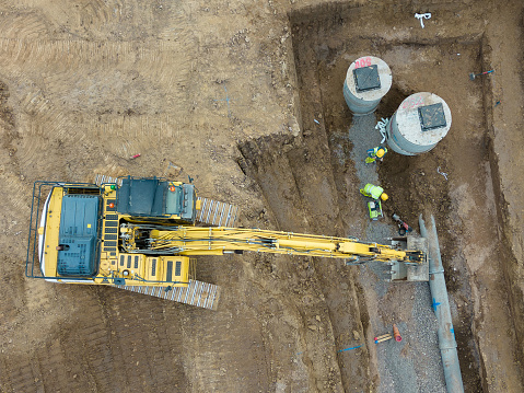 Aerial drone view looking down on a housing development construction site in Scotland.  A bulldozer is at work, digging out a drainage trench ahead of new homes starting to be built. Two construction workers in high visibility workwear are working in the trench.