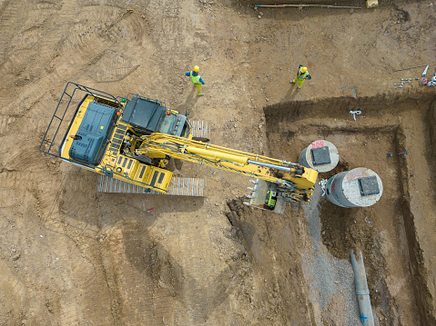 Aerial drone view looking down on a housing development construction site in Scotland.  A bulldozer is at work, digging out a drainage trench ahead of new homes starting to be built. Two construction workers in high visibility workwear are standing nearby.