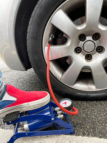 Stock photo showing close-up view of denim jean clad leg of unrecognisable person using a foot pump to inflate a flat car tyre.