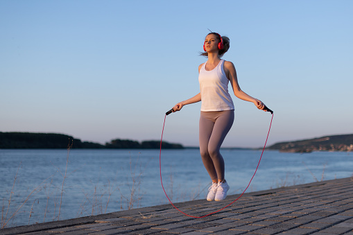 Beautiful young girl in her 20s with blonde hair by the river on a fitness path exercising with jumping rope while listening to music and enjoying the sunset. Photo taken in the golden hour