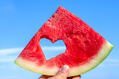 A piece of watermelon with a carved heart inside the hand against the blue sky. Summer mood. Summer background with watermelon. Proper nutrition. Vegetarian food.