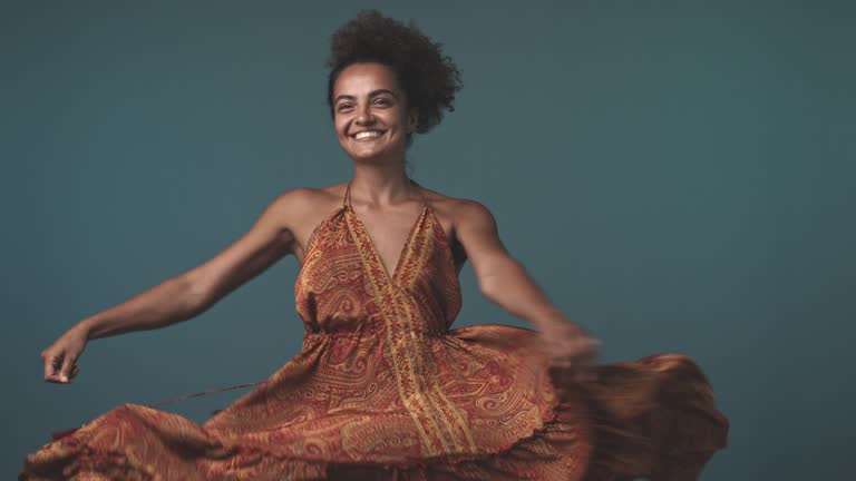 Cheerful young woman spinning and dancing in a flowy dress against teal blue background