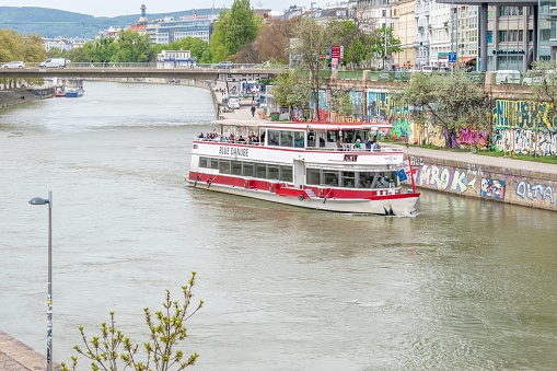 Vienna, Austria – April 28, 2023: A riverboat is seen sailing down a body of water with a graffiti-covered wall and buildings along both sides of the shore in Vienna, Austria