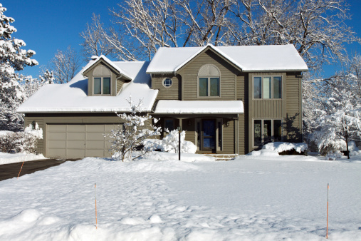 Color DSLR stock picture of green and grey suburban colonial style house covered in bright, white winter snow.  Clear, cold background for home is bright blue sky.  Black top driveway is clear, but lawn is blanketed.  No people in shot but ample copy space for text