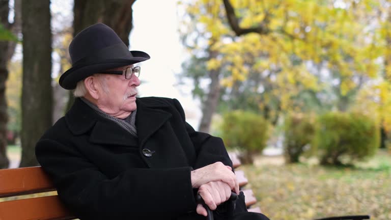 Portrait of an old concentrated grandfather sitting on a bench in autumn season and resting.