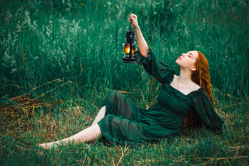 Lonely red hair woman in a green dress with a kerosene lamp is sitting in the grass in the early morning with a kerosene lamp and dreaming. The concept of fairy tale mysticism.