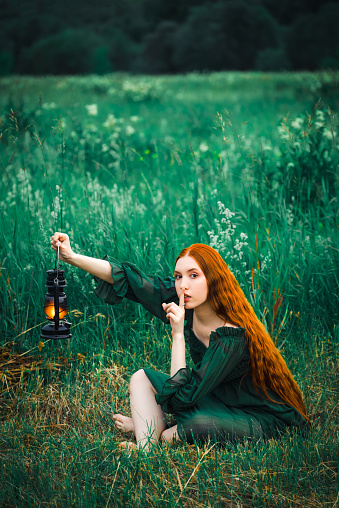 Lonely red hair woman in a green dress with a kerosene lamp is sitting in the grass in the early morning with a kerosene lamp and dreaming. The concept of fairy tale mysticism.