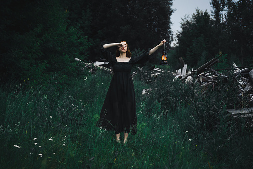 Lonely woman in a black dress with a kerosene lamp in her hands walks through the night forest near the ruins. The concept of fairy tale mysticism.