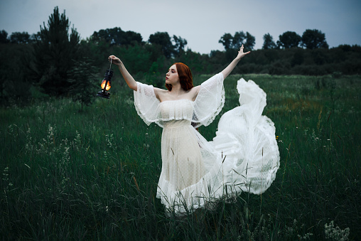 Lonely woman in a white dress with a kerosene lamp in her hands walks through the night field. The concept of fairy tale mysticism.