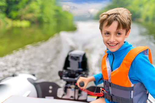 Cute little boy driving the motorboat on a lake surrounded by mountain forest, sunny summer day