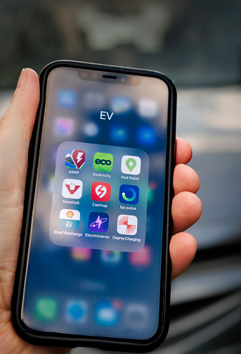 19th August, 2023 - Leeds, UK: A man's hand holding an iPhone, in front of a car. The screen displays a selection of popular apps that are used in the UK for electrical vehicle (EV) charging.