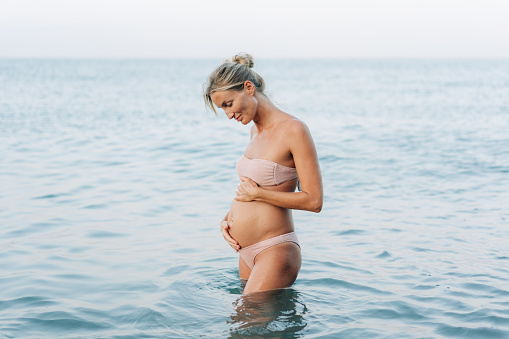 A beautiful pregnant woman stands in the sea and holds her belly. The concept of prenatal care and motherhood.