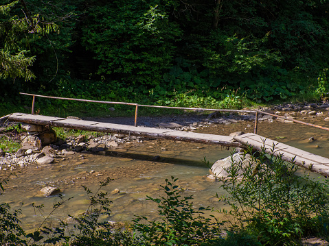 Wooden bridge over river with turquoise waters and rocky riverbed in the forest of Ukraine. Mountain stream flowing through a forest, a small wooden bridge on a hiking trail crossing the river.