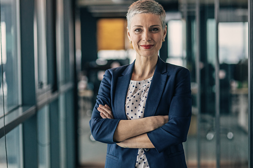 Portrait of mature successful senior woman boss inside office in business suit, businesswoman smiling and looking at camera, standing by window with crossed arms, banker finance woman.