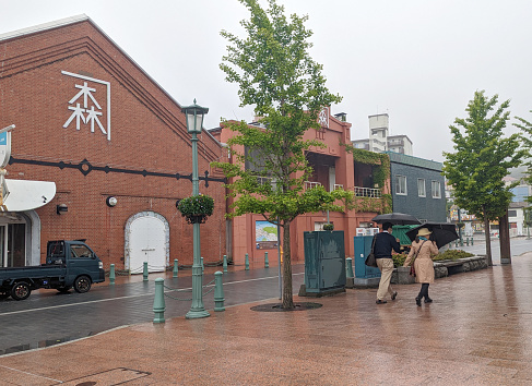 Hakodate, Japan - June 10, 2023: Pedestrians stroll by the Kanemori Red Brick Warehouse in the Hakodate Bay Area. This former warehouse redeveloped as a shopping mall in the port city. Spring morning with rain in Oshima Subprefecture.
