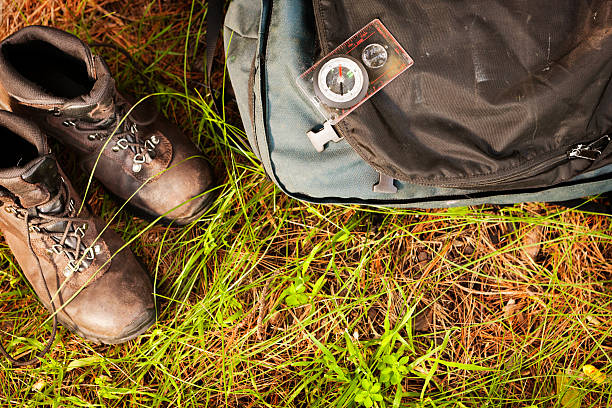 Hiking theme: compass, boots and backpack resting on grass in the countryside. Focus is on the compass.
