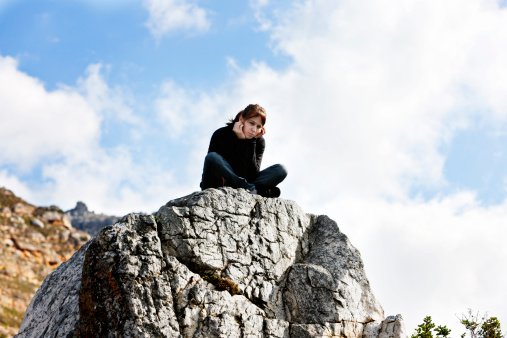 A depressed, grumpy-looking  young woman sits, head in hands, on a rocky clifftop against a blue sky. 